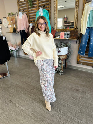 Woman wearing cream turtleneck tunic sweater with floral skirt in boutique clothing store setting.