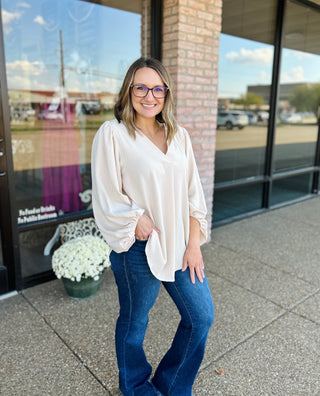 Woman wearing creamy oat balloon sleeve blouse with jeans, standing outside storefront with glasses and smile.