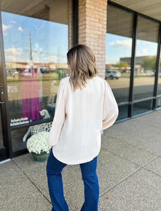 Woman wearing oversized balloon sleeve blouse in cream with jeans, shown from behind outside storefront.