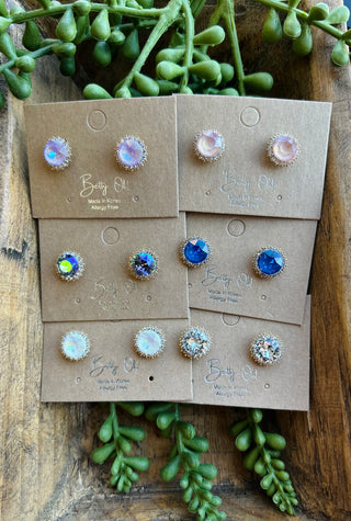 Six pairs of crystal halo stud earrings on branded cards arranged on wooden surface with greenery.