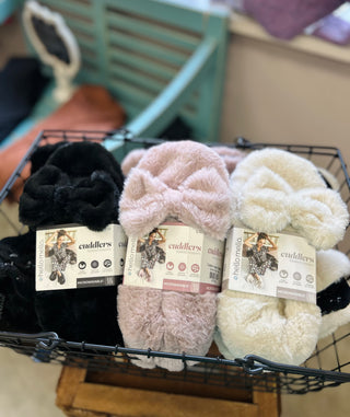 Three pairs of fuzzy heated slippers in a wire basket with product tags attached.