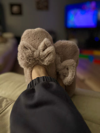 Person wearing cozy fuzzy slippers with bow detail while relaxing on couch watching TV