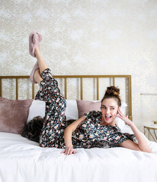 Woman wearing floral pajamas relaxing on bed with cozy heated slippers, smiling in elegant bedroom.
