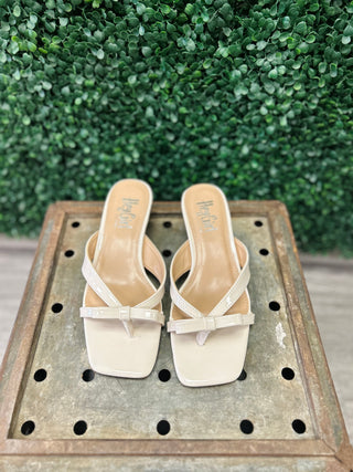 Ivory patent leather sandals with strappy design displayed on wooden surface against green foliage background
