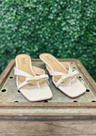 Corkys Dainty ivory patent sandals with bow detail on wooden surface against green foliage background.