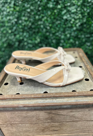 Ivory patent leather sandals with bow detail and low heels displayed on rustic wooden surface.