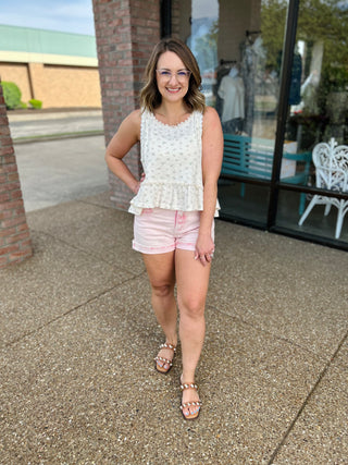 Woman wearing white tiered tank top with pink shorts and sandals, standing on sidewalk outdoors.