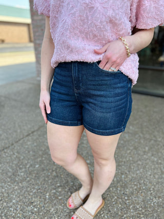 Woman wearing dark blue high-rise denim shorts with pocket details and pink textured top.