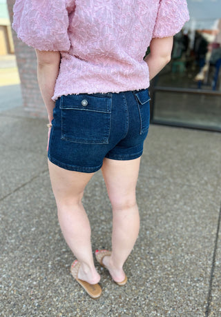 Woman wearing dark blue high-rise denim shorts with pocket details and pink textured top.