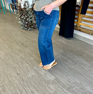 Person wearing dark blue wide leg jeans with hand in pocket, standing on wood floor.