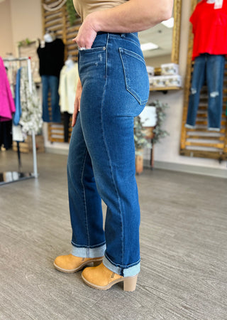 Woman wearing dark wash high-rise straight leg jeans with cuffed hem and tan block heel shoes in clothing store.