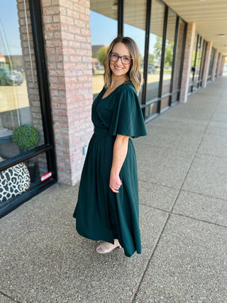Woman wearing a dark green flutter sleeve wrap midi dress standing on sidewalk in front of storefronts.