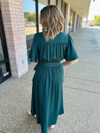 Woman wearing teal flutter sleeve wrap midi dress with belt, walking on sidewalk outside storefront.