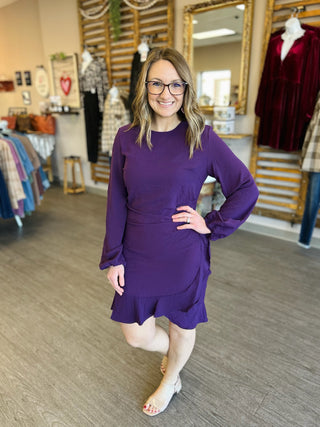 Woman wearing purple long-sleeve ruffle dress with glasses, standing in clothing boutique with one hand on hip.