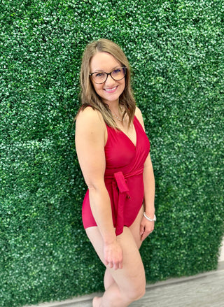 Woman wearing red one-piece swimsuit with tie waist standing against green foliage backdrop