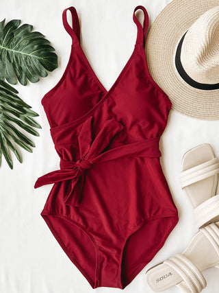 Red one-piece swimsuit with tie waist detail styled with straw hat and sandals on white background.
