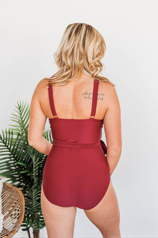 Woman wearing red one-piece swimsuit with tie waist detail, shown from behind in studio setting.