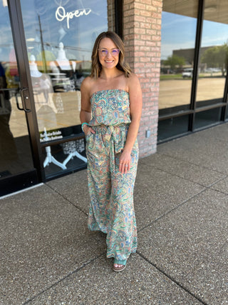 Woman wearing strapless paisley jumpsuit standing outside storefront with glass windows and brick column