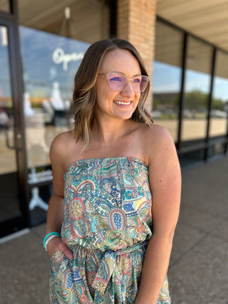Woman wearing strapless paisley jumpsuit with colorful bohemian print, standing outdoors in sunglasses smiling.
