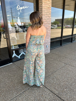 Woman wearing strapless paisley jumpsuit with wide legs, standing outside storefront with "Open" sign