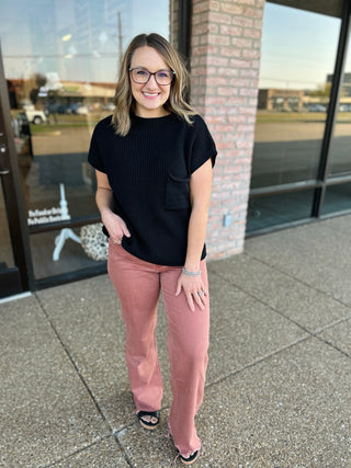 Woman wearing black ribbed sweater top with pink pants, standing outside modern building with brick accents.
