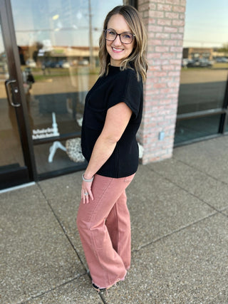Woman wearing black ribbed sweater top with pink wide-leg pants, standing outdoors smiling.