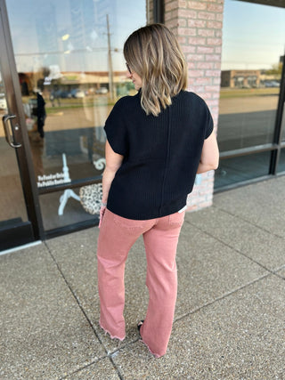 Woman wearing black knit sweater top with pink pants, shown from behind on sidewalk.