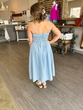 Woman wearing light blue denim midi dress with smocked bodice and spaghetti straps in boutique setting.