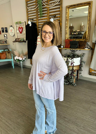 Woman wearing relaxed lavender long-sleeve top with jeans in stylish boutique setting with decorative mirror and plants.