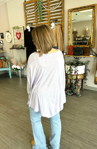 Woman wearing oversized lavender top and jeans standing in boutique with rustic decor and clothing displays.