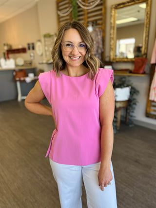 Woman wearing bright pink sleeveless top with ruffle shoulders and white pants in casual indoor setting.