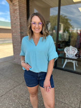 Woman wearing light blue v-neck blouse with denim shorts, standing outdoors smiling at camera.