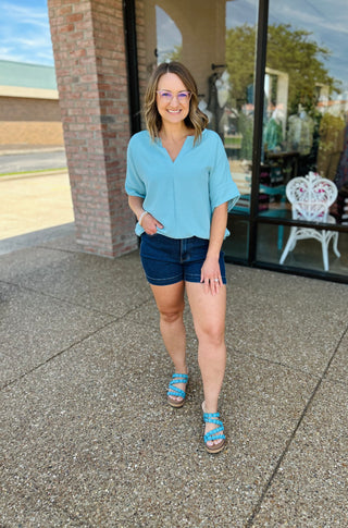 Woman wearing light blue v-neck blouse with denim shorts and sandals, smiling outdoors.