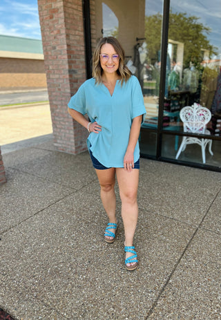 Woman wearing oversized v-neck blouse with short sleeves and denim shorts, standing outside storefront.
