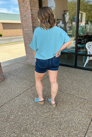 Woman wearing dusty teal v-neck blouse with denim shorts, shown from behind in casual outdoor setting.