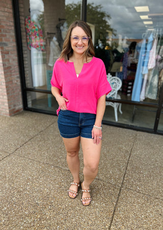 Woman wearing bright pink v-neck blouse with denim shorts, standing outside boutique storefront.