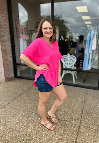 Woman wearing bright pink v-neck blouse with denim shorts, standing outside storefront window.