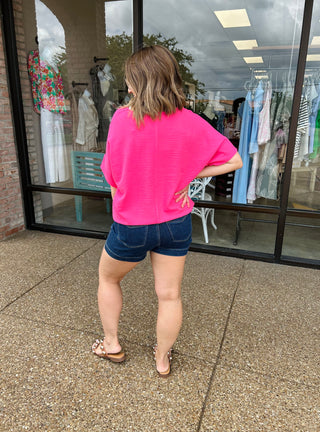 Woman wearing bright pink oversized v-neck blouse with denim shorts, standing outside clothing boutique storefront.