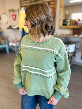 Woman wearing green striped pullover sweatshirt with decorative bow details in casual indoor setting.