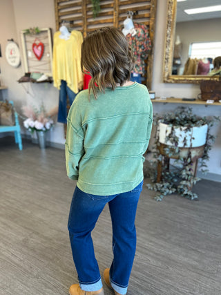 Woman wearing sage green pullover sweatshirt with blue jeans in boutique clothing store setting.