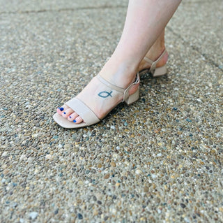 Woman wearing beige block heel sandals with ankle strap on textured concrete surface, blue pedicure visible.