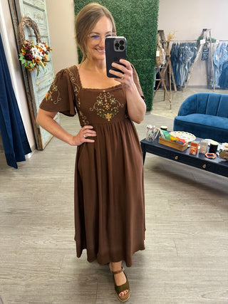 Woman wearing an embroidered midi dress with bell sleeves, square neckline, and floral cross-stitch detailing.