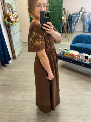 Woman wearing an embroidered floral midi dress with bell sleeves, taking a mirror selfie in a boutique.