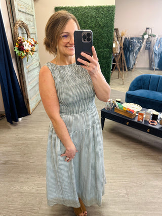 Woman wearing a sleeveless gingham midi dress with smocked waist, taking a mirror selfie in a boutique.