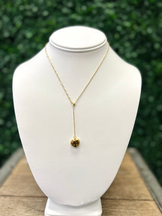 Gold heart pendant necklace displayed on white jewelry bust against green foliage background.