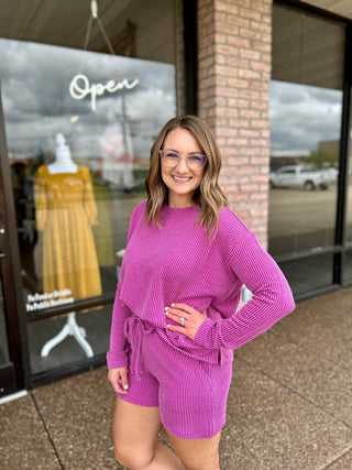 Woman wearing a matching ribbed long-sleeve top and shorts set, posing outside a boutique.