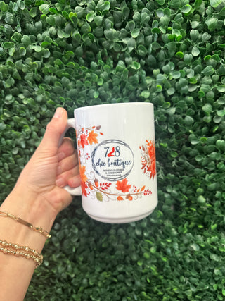 White ceramic mug featuring autumn leaves design and 728 Chic Boutique logo, held against green foliage backdrop.