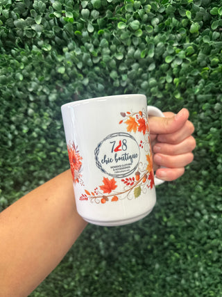 White ceramic mug featuring fall foliage design and 728 Chic Boutique logo, held against green wall.
