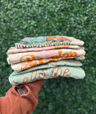 Hand holding stack of colorful graphic tees and sweatshirts with fall-themed designs against green foliage background.