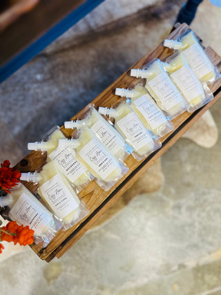 Squeezable wax melts in clear pouches with white labels arranged on wooden display tray with flowers.